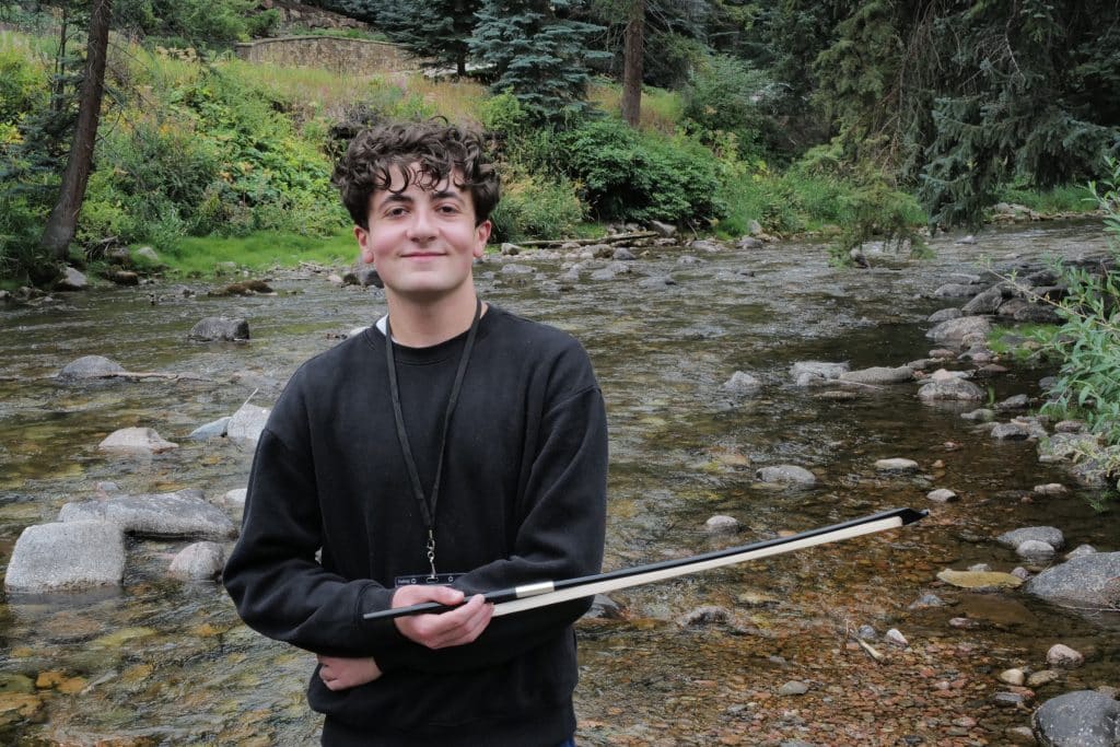 A Vail Jazz Alumni with curly hair wearing a black sweater stands by a rocky stream, holding a violin bow, surrounded by lush green trees and plants.
