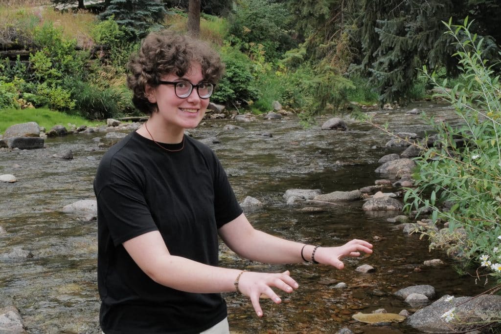 A Vail Jazz Alumni with short curly hair, glasses, and a black shirt stands by a rocky stream, smiling and holding their hands up playfully amid lush green, wooded surroundings.