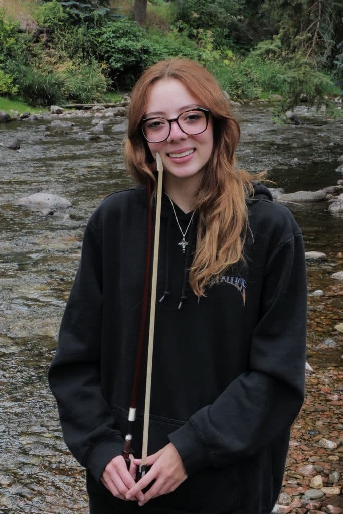 A young woman with long, light brown hair and glasses stands by a rocky stream, smiling and holding a violin bow vertically in front of her face. A proud Vail Jazz Alumni, she wears a black hoodie and is surrounded by greenery and water.