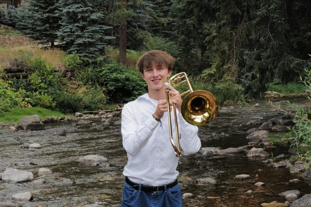 A young Vail Jazz Alumni in a white shirt and jeans stands in a rocky creek holding a brass trombone, smiling at the camera. Tall evergreen trees and lush greenery fill the background.
