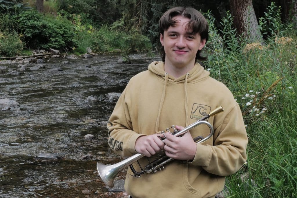 A young person in a tan hoodie stands by a rocky stream, holding a trumpet and smiling, surrounded by green trees and tall grass—a proud Vail Jazz Alumni enjoying nature.
