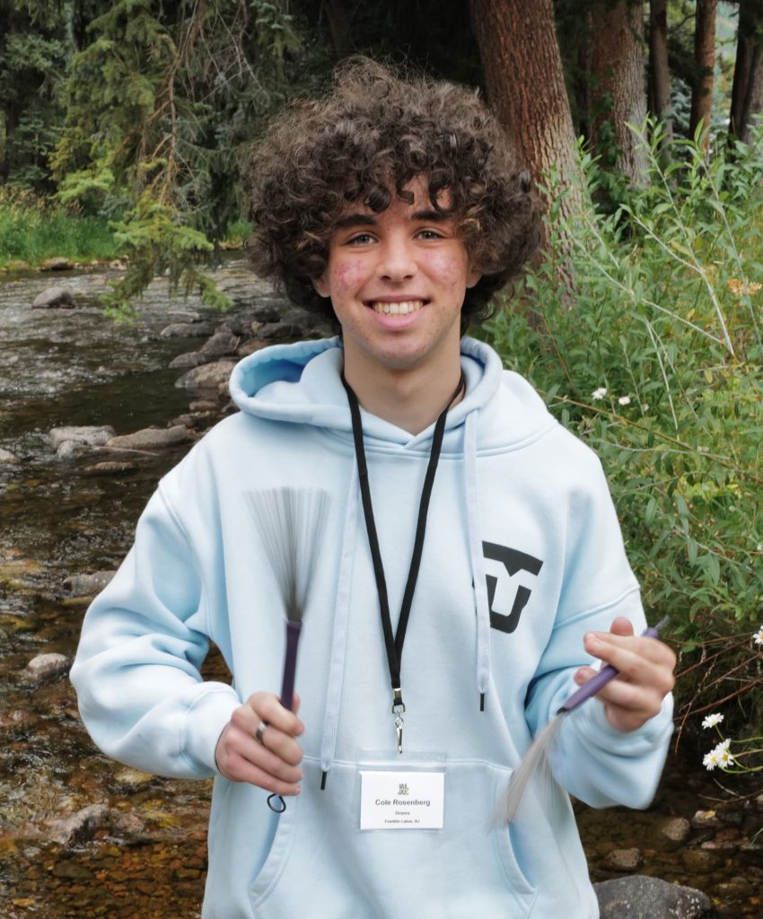 A smiling curly-haired teen, Vail Jazz Alumni, in a light blue hoodie and plaid pants stands by a creek, holding two percussion brushes, with a name badge around their neck and trees in the background.