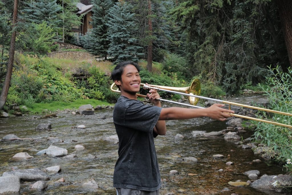A Vail Jazz Alumni stands in a rocky creek surrounded by trees, smiling while playing a brass trombone. A wooden house is visible in the background among the greenery.