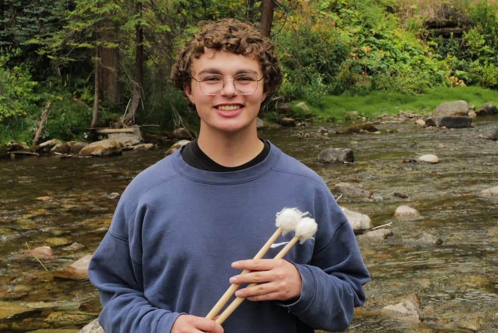 A smiling Vail Jazz Alumni with curly hair and glasses stands by a rocky stream, holding two mallets with white fluffy heads. They wear a blue sweatshirt, surrounded by trees and greenery in the background.