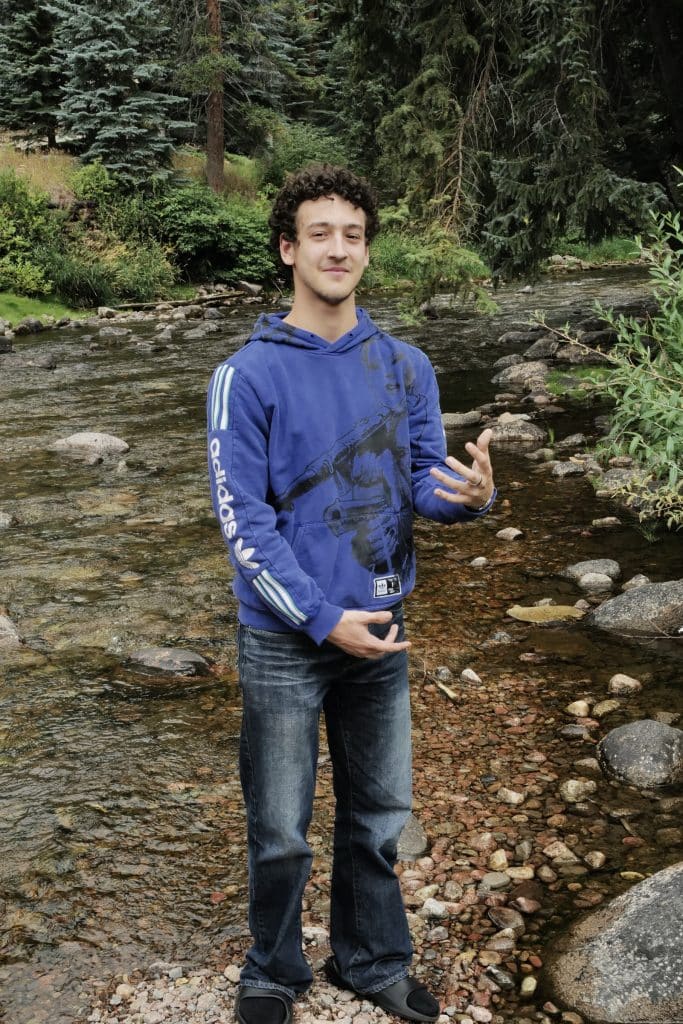 A young man in a blue Adidas hoodie and jeans, smiling and gesturing with his hands, stands on a rocky bank by a shallow stream surrounded by lush greenery—a proud Vail Jazz Alumni enjoying nature.