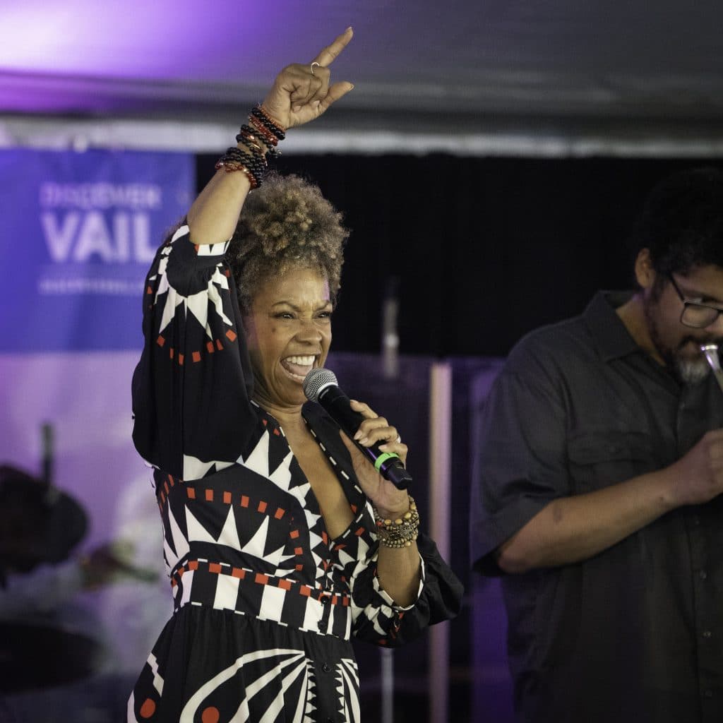 A woman with curly hair sings passionately into a microphone, raising one arm with her finger pointed upward. She wears a black dress with red and white geometric patterns and multiple bracelets. A man stands beside her in the background.