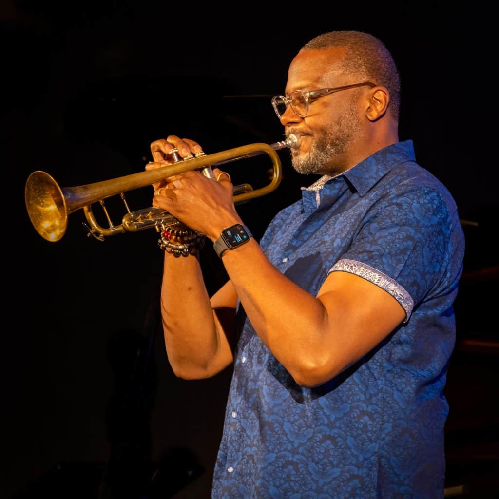 A man wearing glasses and a blue patterned shirt plays a trumpet against a dark background, with a focused expression and an Apple Watch on his wrist.