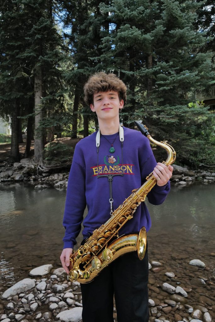 A young Vail Jazz Alumni in a purple Branson sweatshirt holds a saxophone while standing on rocks by a creek, with tall trees in the background.